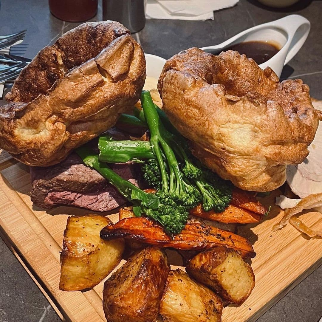 Traditional Sunday roast with Yorkshire puddings, beef slices, roasted potatoes, carrots, and broccolini served on a wooden board at Mad Frans Bar.