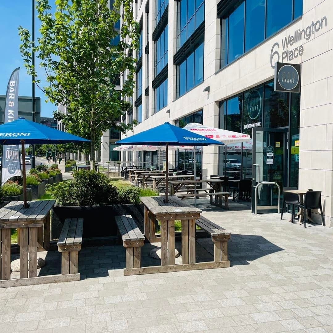 Outdoor seating area with wooden benches and blue umbrellas at Mad Frans Bar, located at 6 Wellington Place on a sunny day.