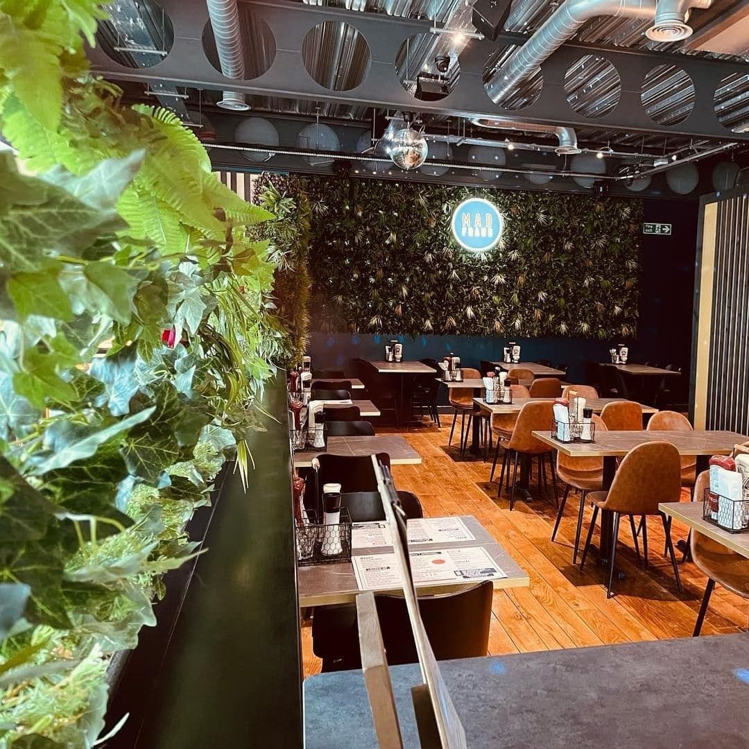 Interior of Mad Frans Bar with tables set for dining, a green plant wall in the background, and the bar's circular logo illuminated on the wall.