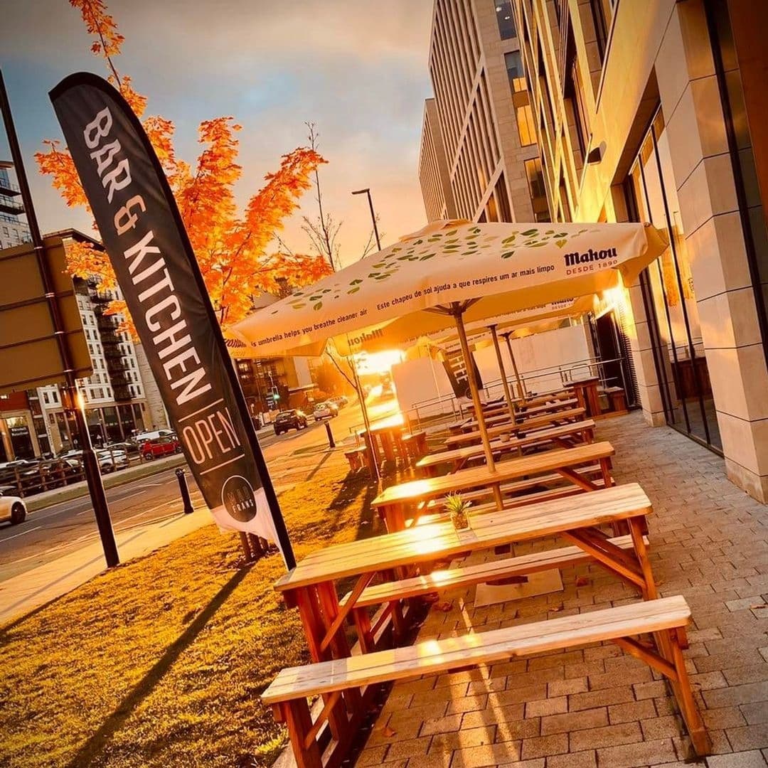 Outdoor seating at Mad Frans Bar with wooden picnic tables, umbrellas, and a 'Bar & Kitchen Open' flag during a sunset.