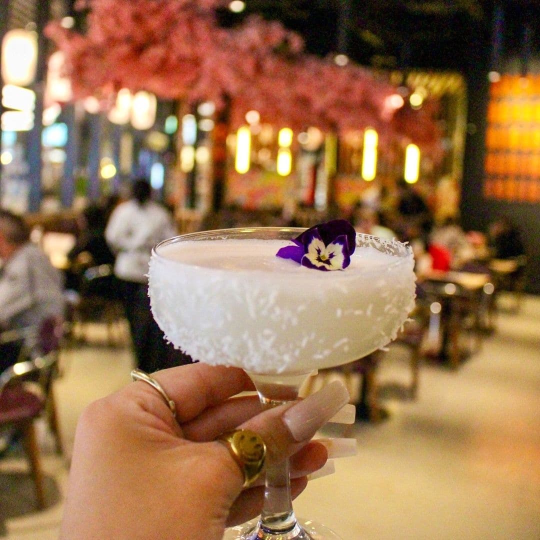 Hand holding a cocktail garnished with a purple flower at Maki & Ramen, with a backdrop of cherry blossom trees and warm ambient lighting.