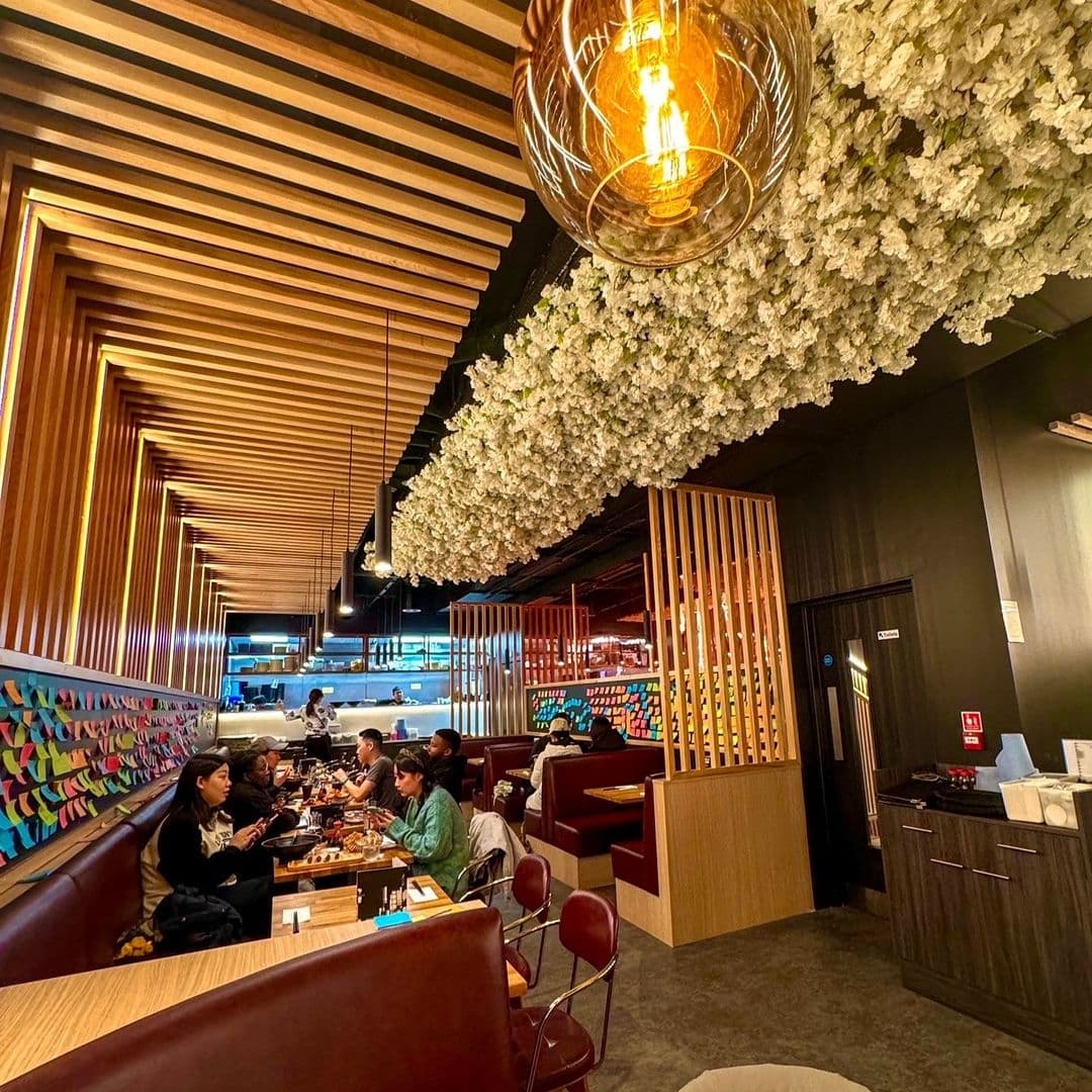 Interior of Maki & Ramen restaurant featuring diners at tables, decorative ceiling with wooden slats and hanging light bulb, and a wall adorned with white flowers.