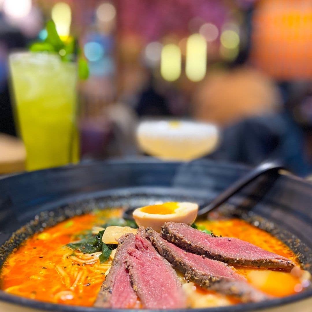 Close-up of a vibrant bowl of ramen with slices of pink-centered beef, half an egg, and greens at Maki & Ramen, with a blurred background featuring a green drink and warm ambient lighting.