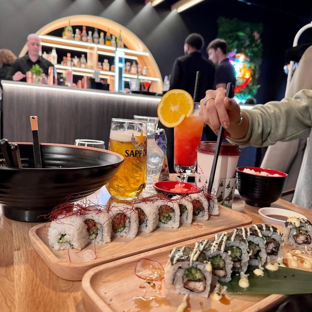 Dining experience at Maki & Ramen featuring a close-up of sushi rolls on a wooden tray, a bowl of ramen, a glass of Sapporo beer, and a fruity cocktail, with a customer in the background using chopsticks.