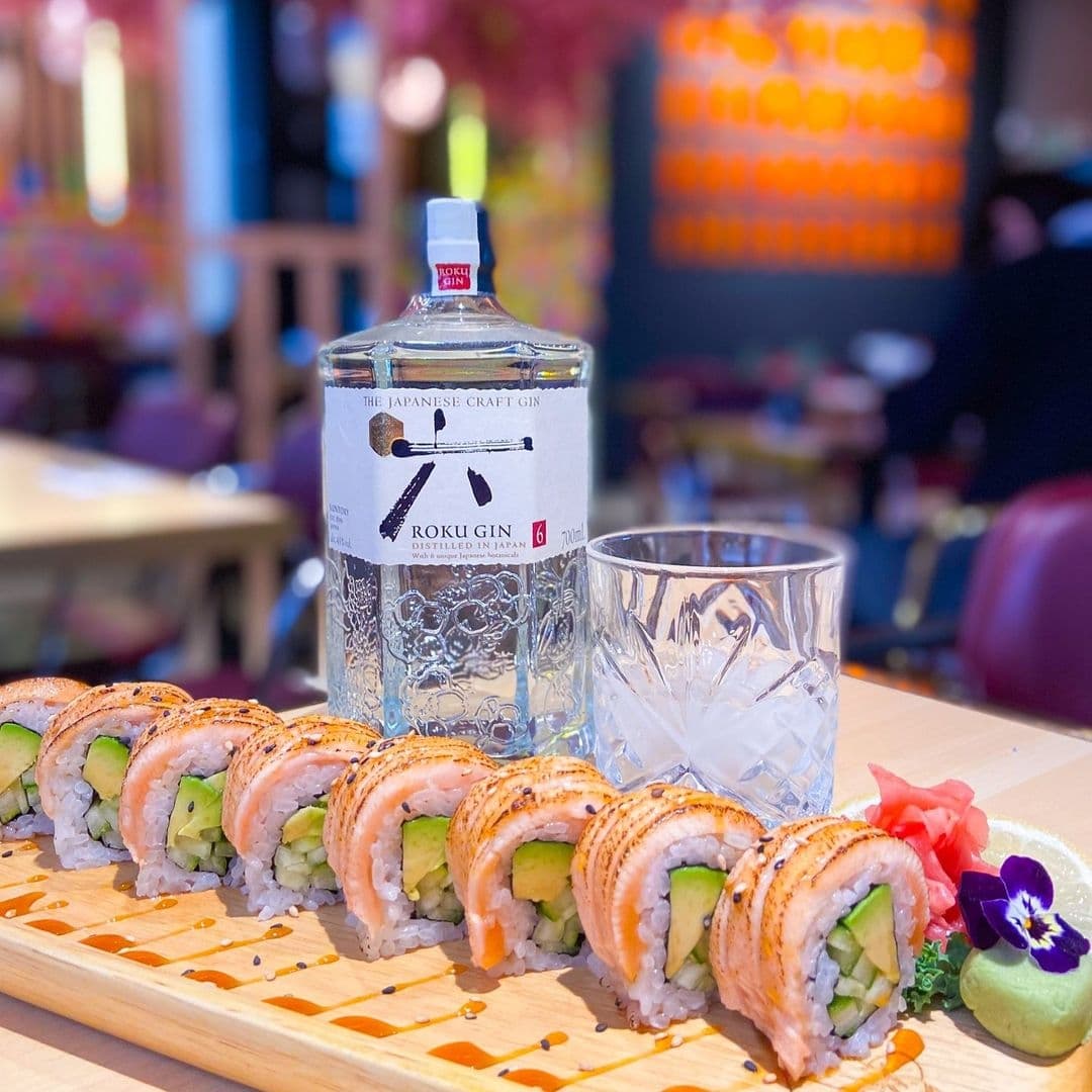 Sushi rolls with salmon on top, cucumber inside, and garnishes, with a bottle of Roku Gin and a glass on a wooden table at Maki & Ramen.