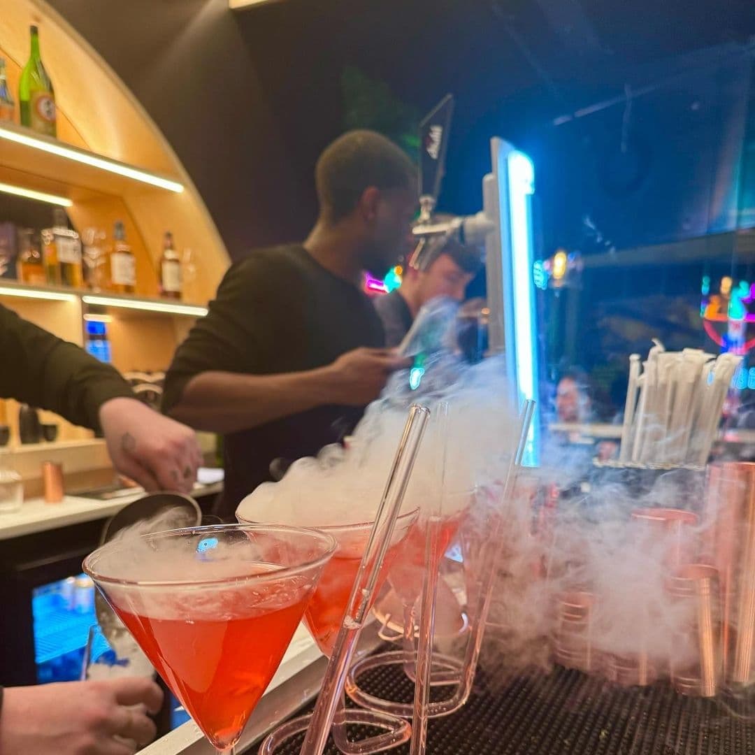 Vibrant atmosphere at Maki & Ramen with a bartender preparing drinks, featuring a red cocktail with dramatic dry ice smoke effect on the bar counter.