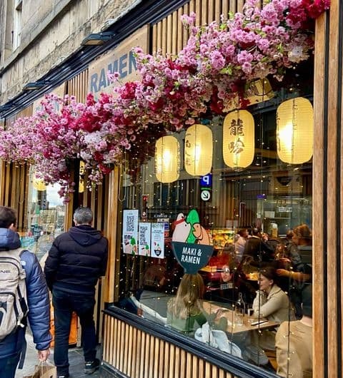 Exterior view of Maki & Ramen restaurant with vibrant pink flowers above the window, traditional lanterns, and patrons dining inside.