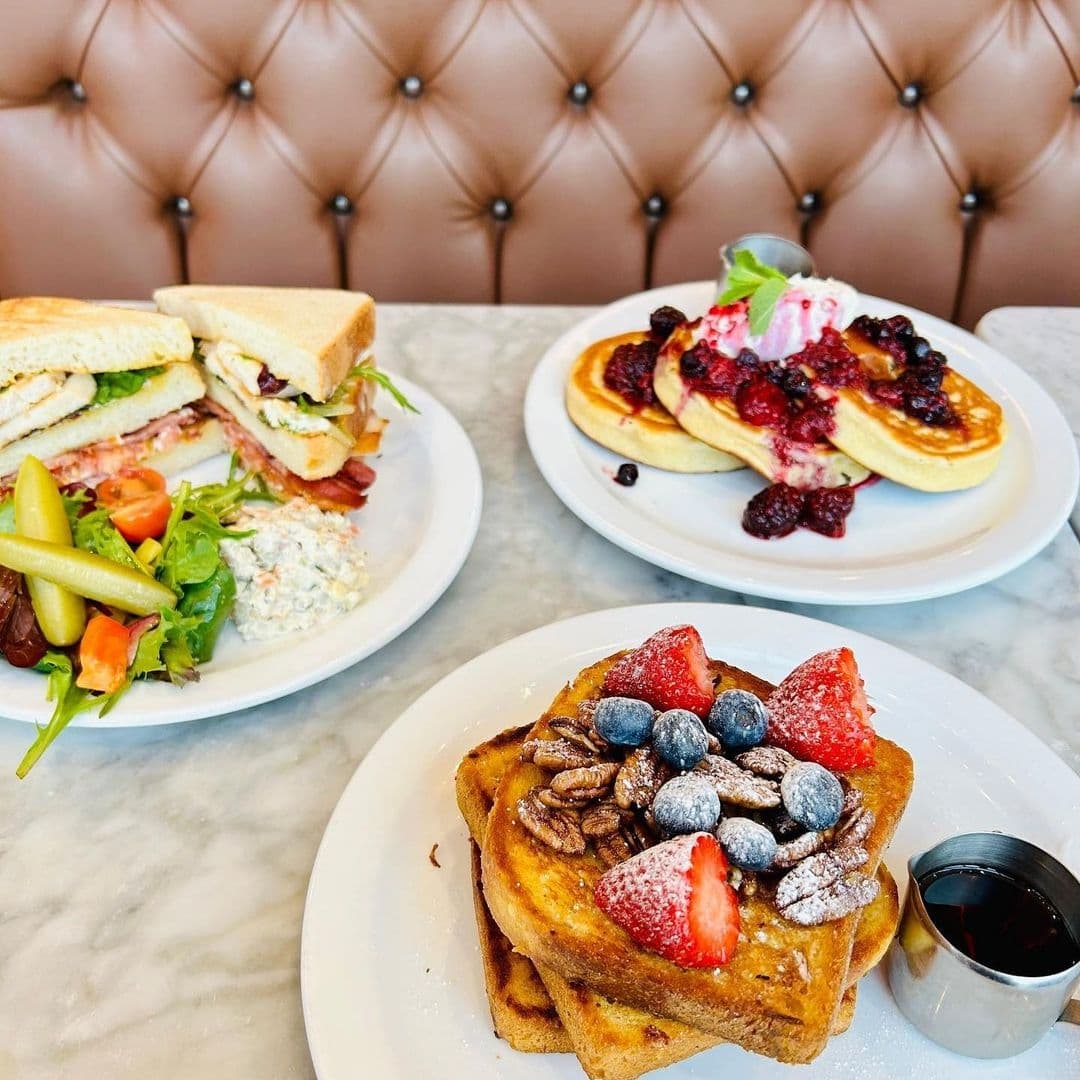 Delicious breakfast spread at Moose Coffee featuring a club sandwich, pancakes with berry compote and whipped cream, and French toast topped with strawberries and blueberries, served with syrup on the side.