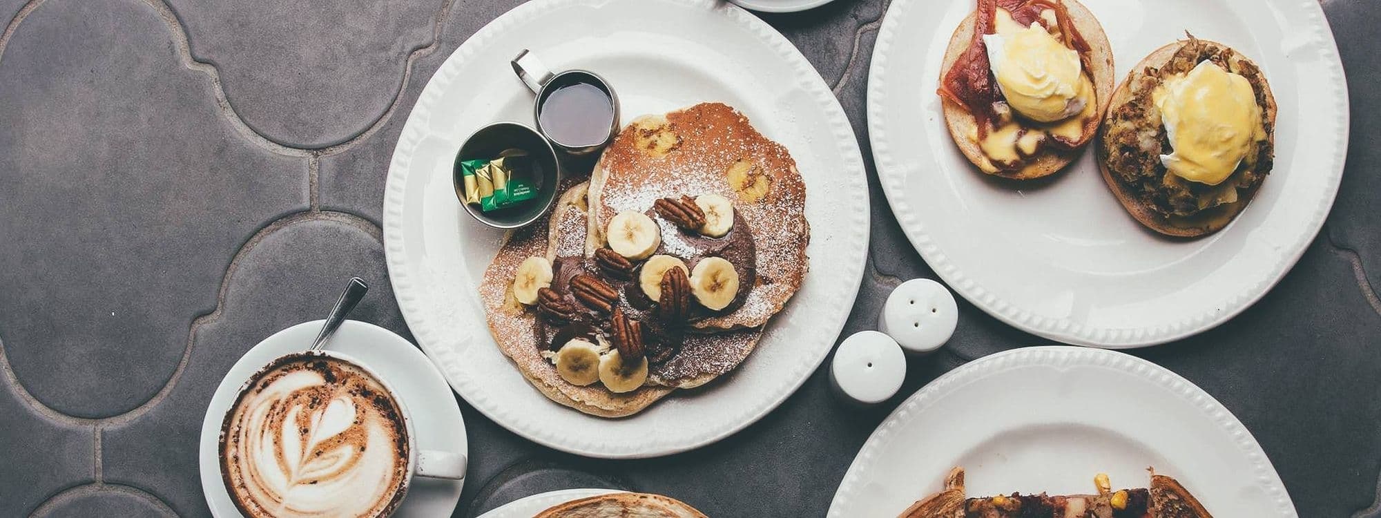 Delicious breakfast spread at Moose Coffee featuring pancakes with bananas and pecans, eggs benedict on English muffins with bacon, and a cappuccino with latte art, all served on white plates on a gray tabletop.