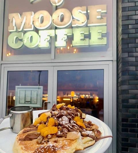 A plate of pancakes topped with chocolate sauce and dried fruit in the foreground with the illuminated sign of Moose Coffee in the background during twilight.