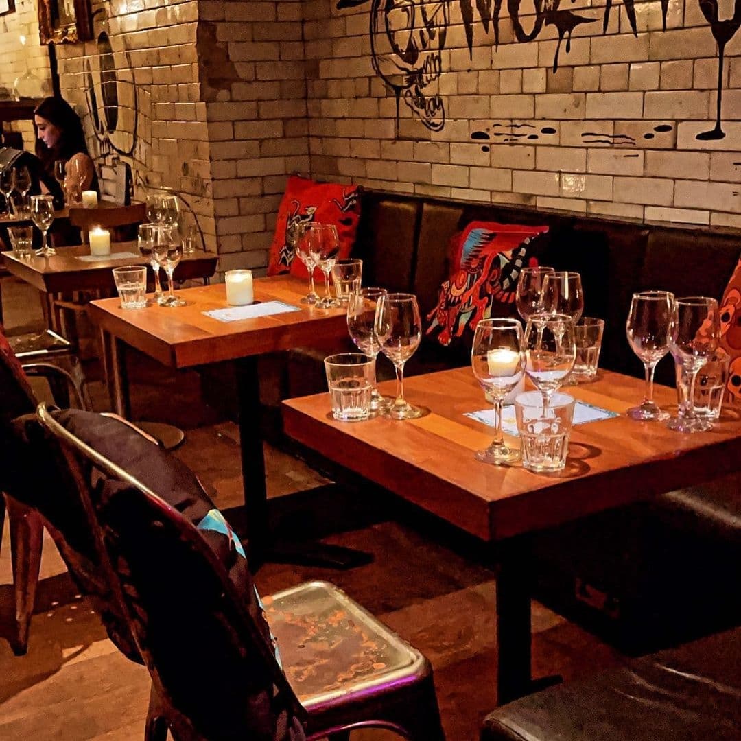 Interior of Neon Cactus showing a cozy dining setup with lit candles on wooden tables, wine glasses ready for guests, and vibrant cushioned seats against a brick wall with artistic graffiti.