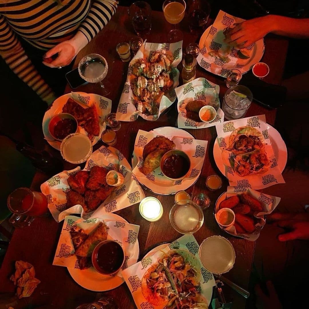 Diners enjoying a variety of dishes and drinks at Neon Cactus, with plates of tacos, nachos, and wings spread across a wooden table, illuminated by warm, ambient lighting.