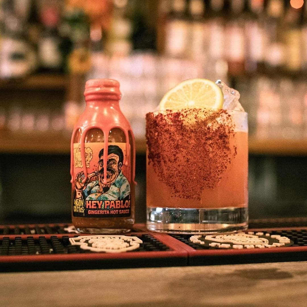 A vibrant cocktail with a lemon slice and a spicy rim, next to a bottle of Neon Cactus Hey Pablo hot sauce, displayed on a bar counter.