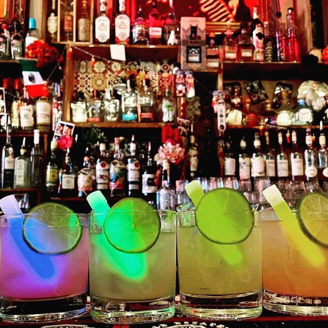 Four colorful cocktails with lime garnishes on a bar counter at Neon Cactus, with a vibrant display of bottles and decorations in the background.
