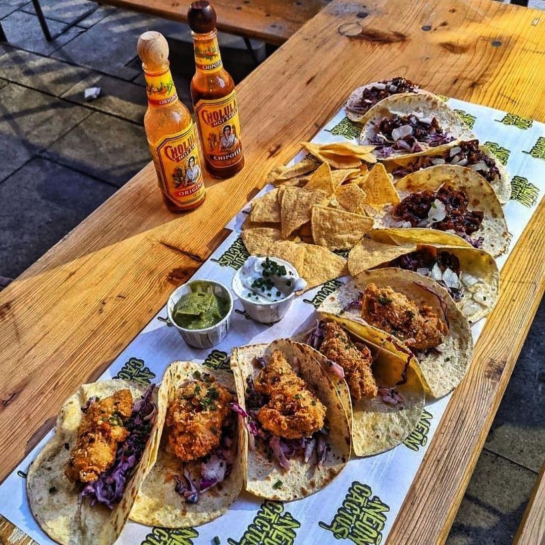 Alt text: "Delicious array of Mexican cuisine at Neon Cactus, featuring crispy fish tacos with purple cabbage slaw, alongside tortilla chips, guacamole, and sour cream, complemented by bottles of Cholula hot sauce."