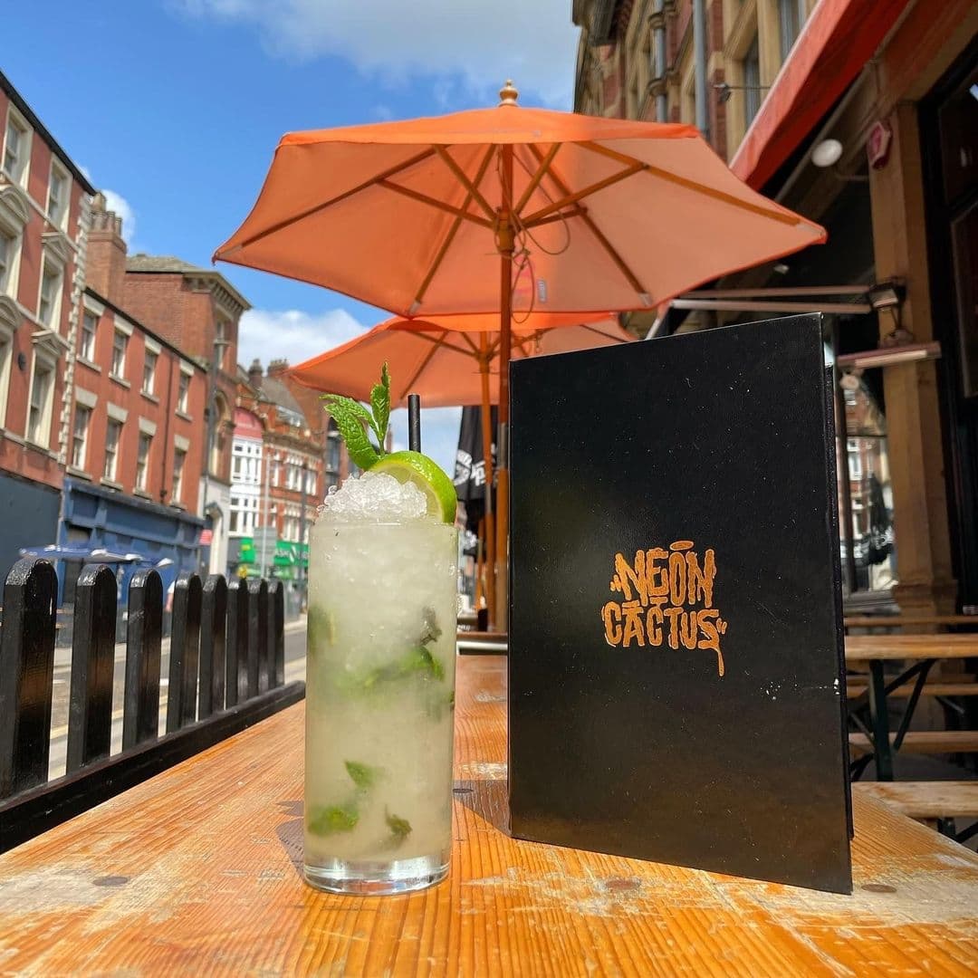 A refreshing mojito with lime and mint on a wooden table at Neon Cactus, with a black menu featuring the location's logo, and a vibrant orange umbrella in the background on a sunny day.