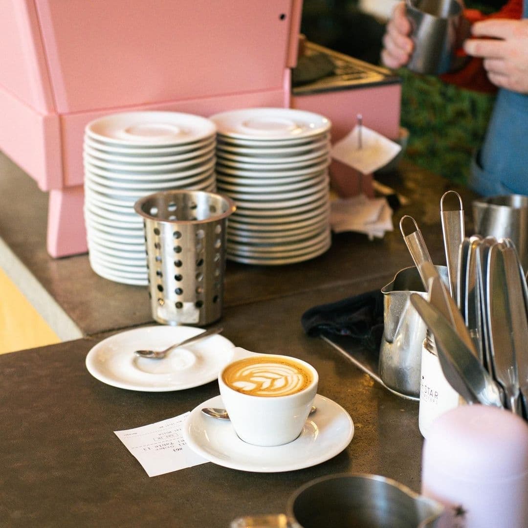 A freshly brewed latte with artful foam design sits on a counter at North Star Coffee Shop, surrounded by stacks of clean white plates and utensils, with a barista in the background.