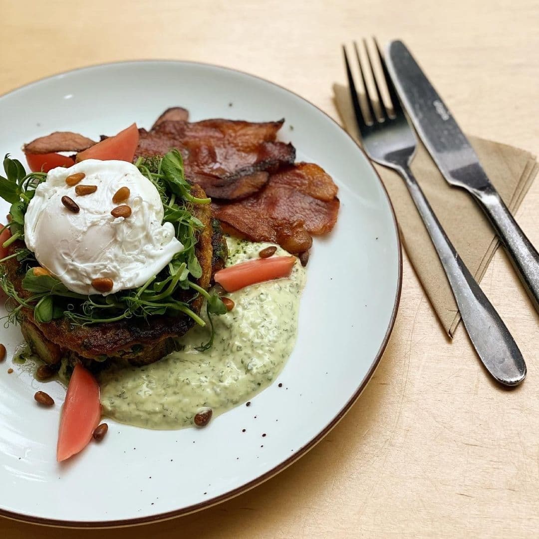 Gourmet breakfast at North Star Coffee Shop featuring a poached egg atop a vegetable hash, garnished with microgreens and seeds, served with crispy bacon, fresh tomato slices, and a creamy herb sauce, accompanied by silverware on a wooden table.