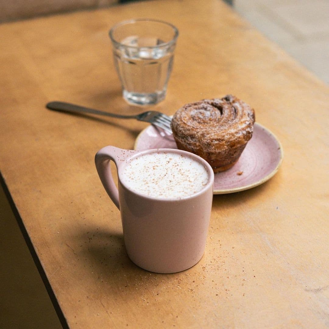 A cozy setting at North Star Coffee Shop featuring a frothy cappuccino in a pink mug, a cinnamon roll on a pink plate, and a glass of water on a wooden table.