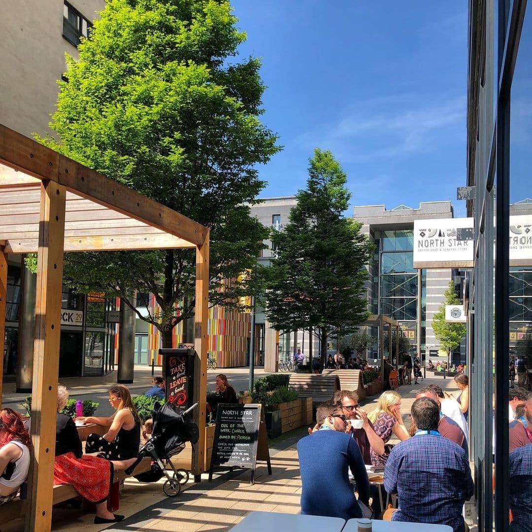Sunny outdoor seating at North Star Coffee Shop with patrons enjoying drinks and conversation, surrounded by green trees and urban architecture.