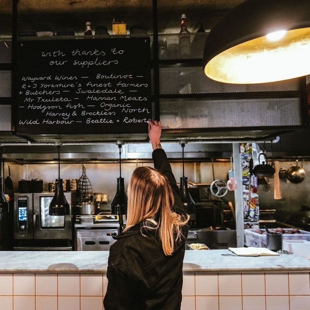 Woman writing on a blackboard listing suppliers at Ox Club at Headrow House, with a kitchen counter and cooking equipment in the background.