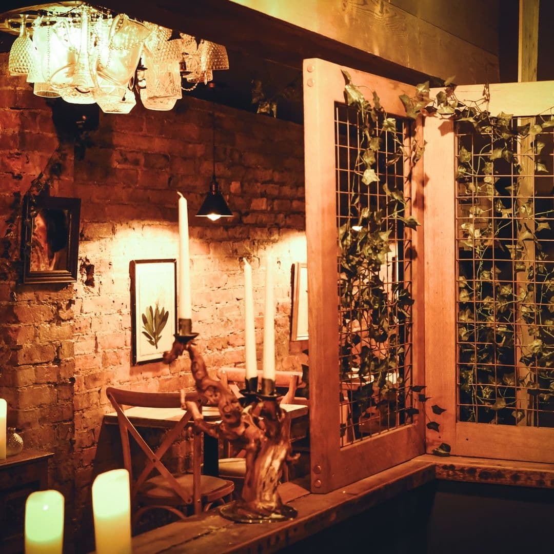 Cozy dining corner at Phranakhon with rustic brick wall, vintage chandelier, and wooden furniture adorned with candles and greenery.