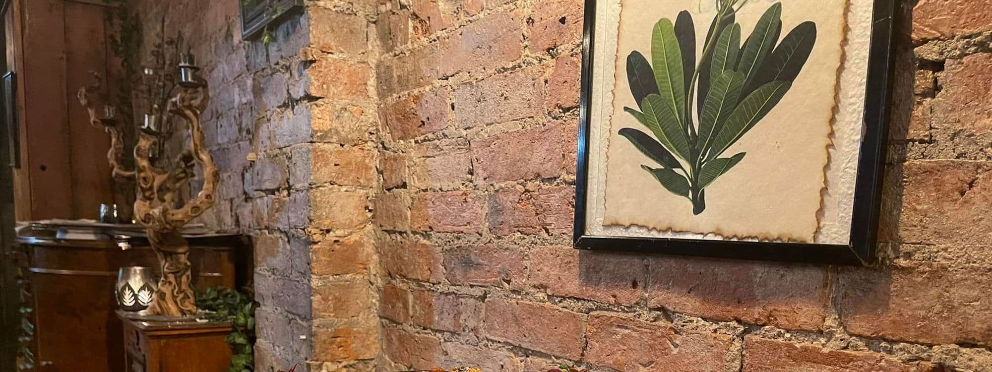 "Interior wall of a Phranakhon venue featuring exposed brickwork and a framed botanical illustration, with a rustic candelabra on a wooden surface."