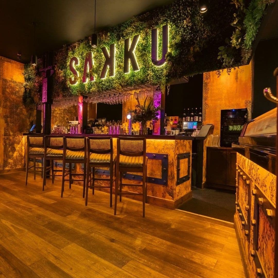 Interior of Sakku bar with illuminated 'SAXKU' sign over a greenery-adorned wall, rustic bar counter with stools, and warm ambient lighting.