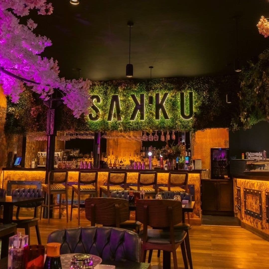 Interior of Sakku bar with illuminated green 'SAKU' sign above the bar counter, surrounded by decorative greenery and pink blossoms.