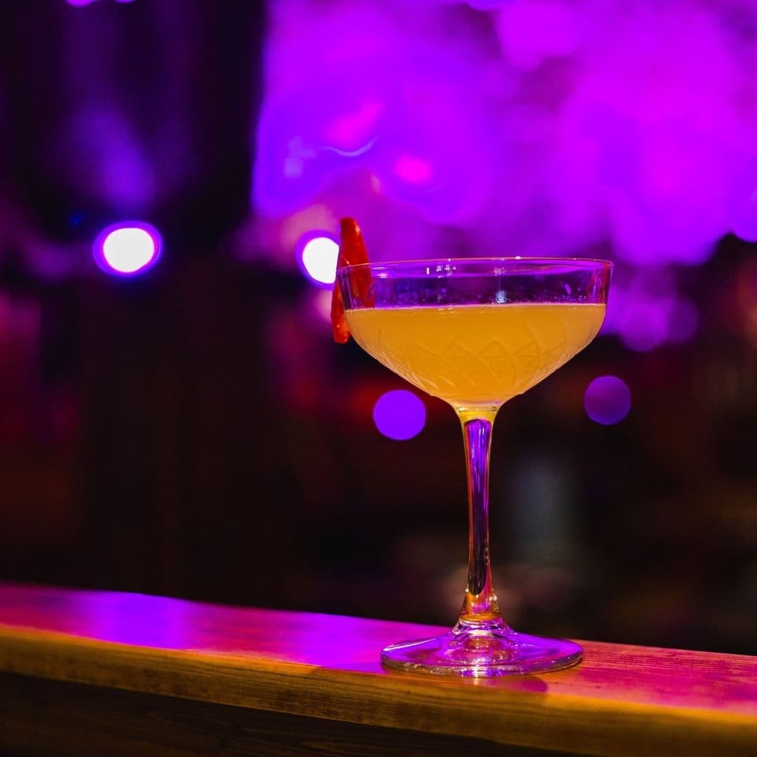 Cocktail glass with a yellow drink garnished with an orange peel at Sakku, with purple ambient lighting in the background.