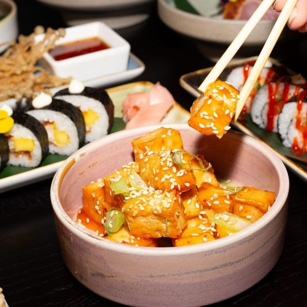 Alt text: "Delicious assortment of sushi and tofu dish at Sakku, with chopsticks holding a piece of tofu topped with sesame seeds and sauce."
