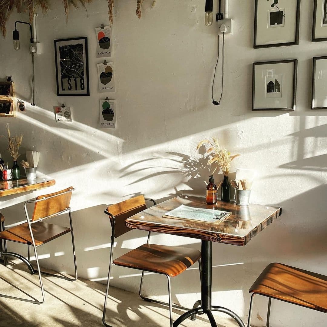 Interior of Slow Rise Bakery with sunlight streaming through a window, casting shadows on a white wall with framed pictures, beside a cozy seating area with wooden tables and chairs.