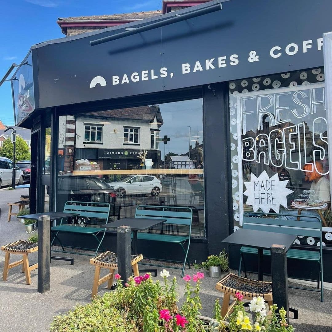 Exterior view of Slow Rise Bakery featuring a black storefront with signage "BAGELS, BAKES & COFFEE," outdoor seating with green metal chairs and wooden stools, and a window display with a sign reading "FRESH BAGELS MADE HERE." Bright flowers in the foreground add a welcoming touch.