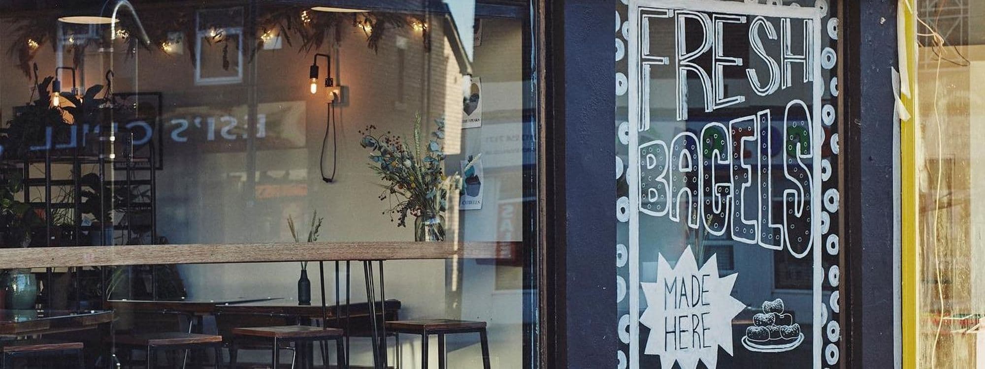 Exterior view of Slow Rise Bakery with a window display featuring a sign reading "FRESH BAGELS MADE HERE" and a cozy interior with plants and pendant lighting visible through the glass.