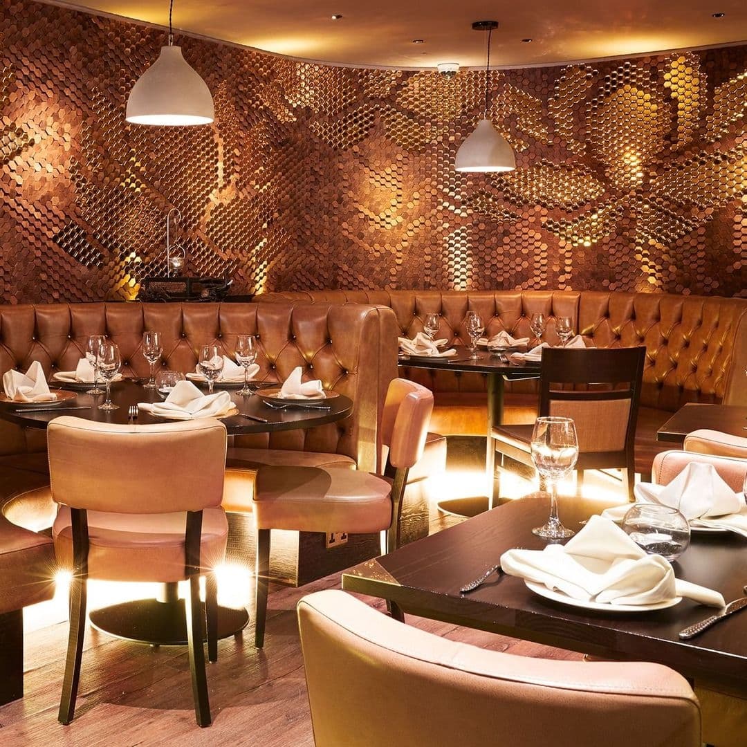 Elegant dining area at Sukhothai Chapel Allerton featuring plush tan booths, wooden tables set for service, and unique copper-toned wall decor, illuminated by soft pendant lighting.