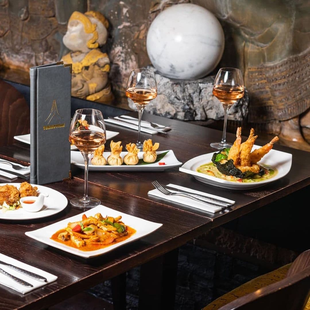 Elegant dining setup at Sukhothai Chapel Allerton featuring Thai cuisine with dishes and rose wine glasses on a wooden table, accented by a menu and traditional Asian decor in the background.