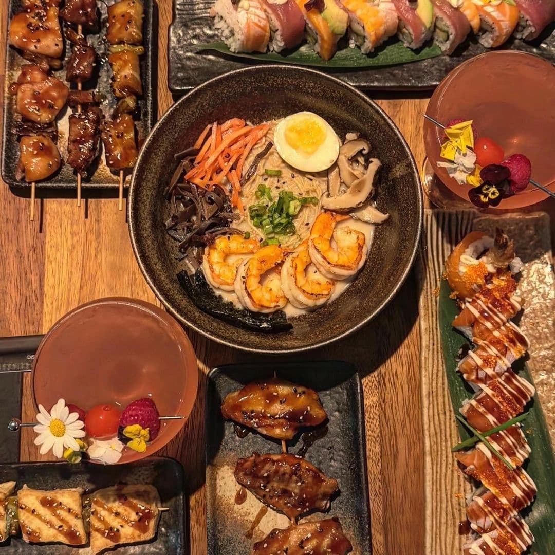 Assorted Japanese cuisine at TADA featuring yakitori, ramen with shrimp and egg, sushi, and a cocktail garnished with a flower.