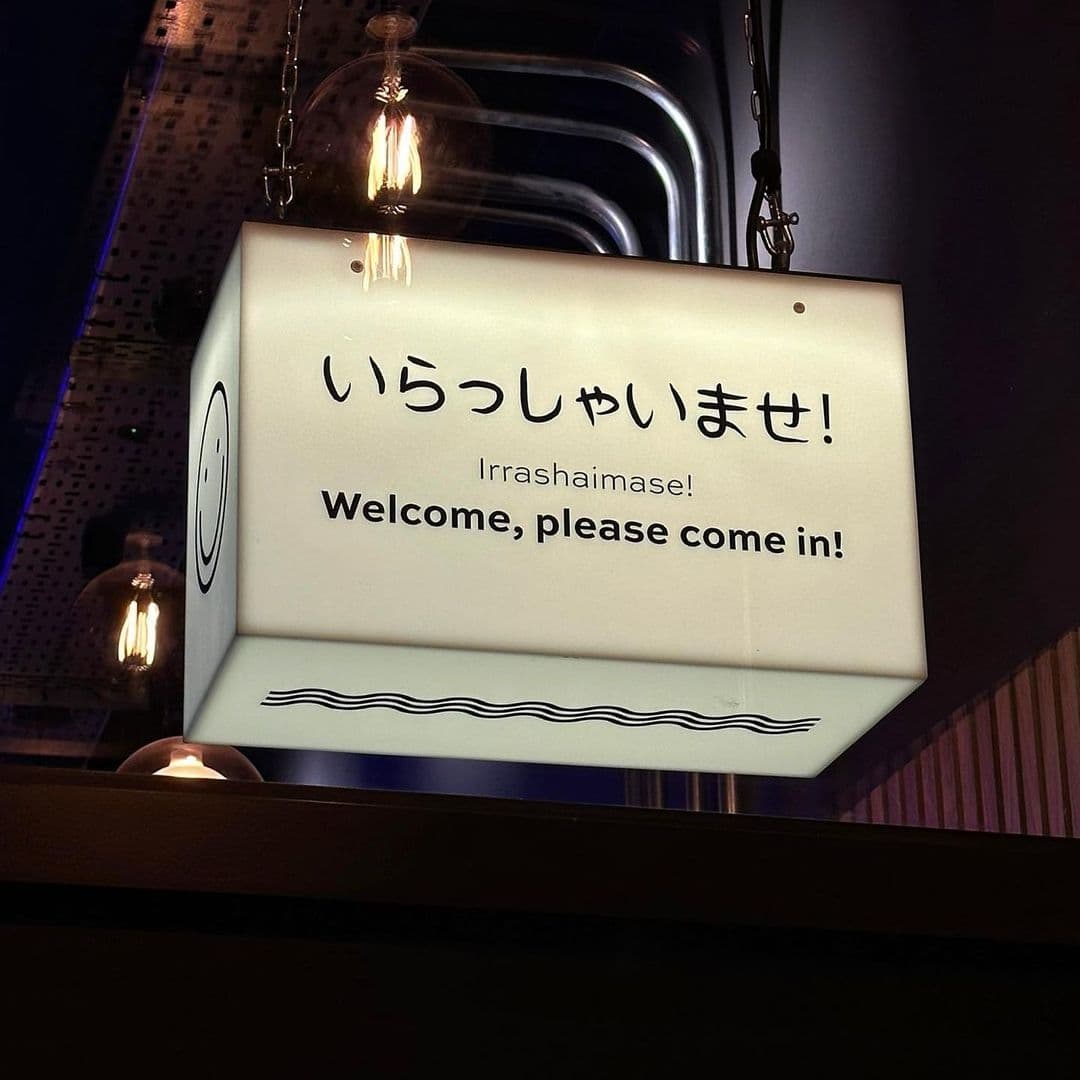Illuminated sign with "いらっしゃいませ! Irrashaimase! Welcome, please come in!" written in Japanese and English at TADA, inviting visitors to enter.