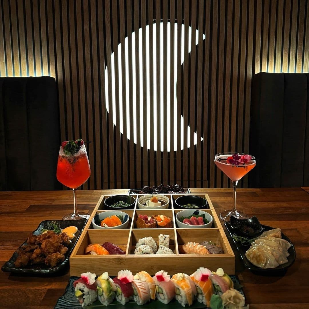 Assorted sushi and Japanese dishes with colorful cocktails at TADA restaurant, featuring elegant wooden decor and ambient lighting.