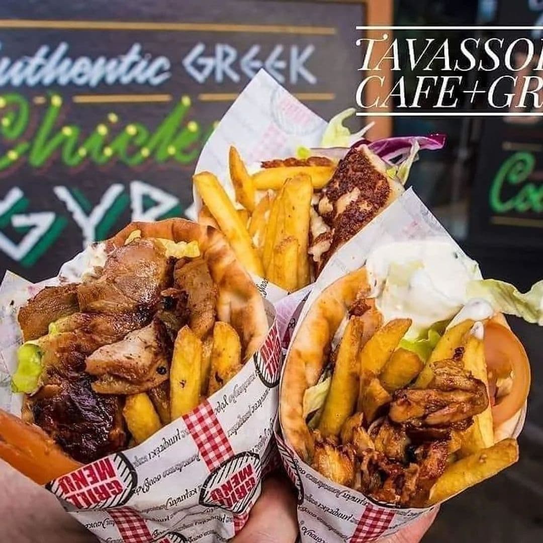 Close-up of two hands holding gyros filled with grilled meat, fries, and sauce from Tavassoli's Cafe and Grill, with a blurred sign in the background.