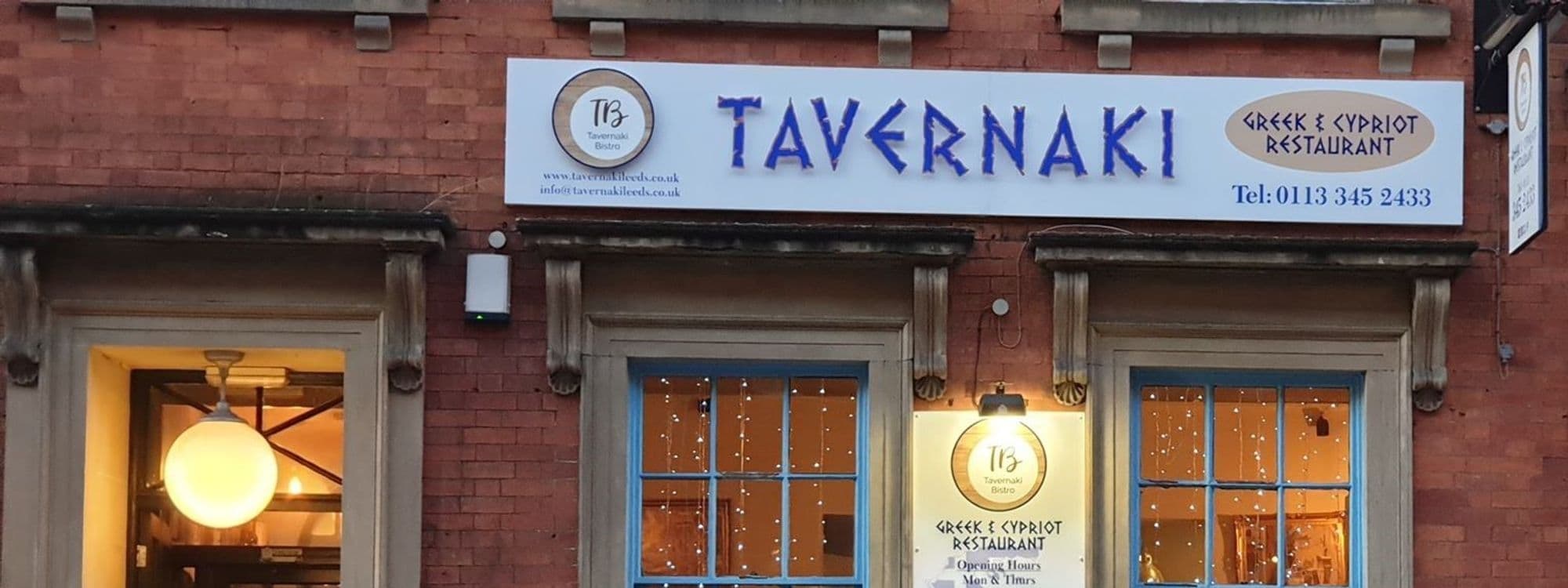 Exterior view of Tavernaki Bistro, showcasing the restaurant's signage with the name "TAVERNAKI" in large blue letters and additional text stating it is a Greek & Cypriot Restaurant, along with contact information. The bistro is housed in a red brick building with large windows illuminated by warm interior lighting.