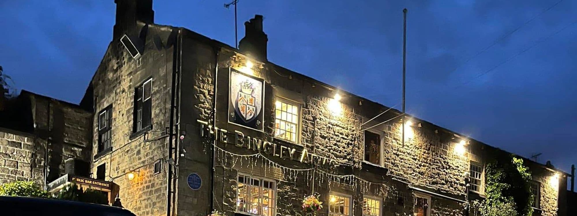 Exterior view of The Bingley Arms at night, showcasing its illuminated sign and stone facade under a dark blue sky.