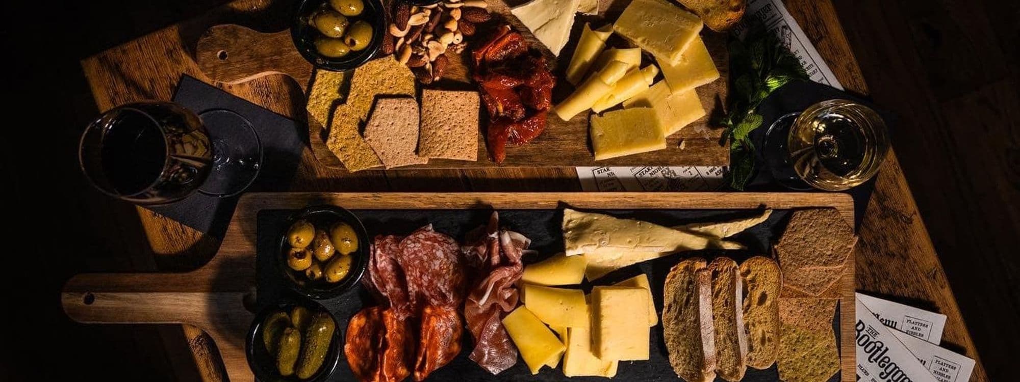Charcuterie board with assorted cheeses, meats, olives, and crackers, accompanied by two glasses of wine, presented on a rustic wooden table at The Bootlegger.