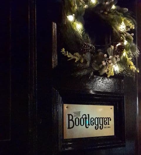 Entrance to The Bootlegger, featuring a dark wooden door with a decorative wreath and a sign with the establishment's name and the year it was established, 2014.
