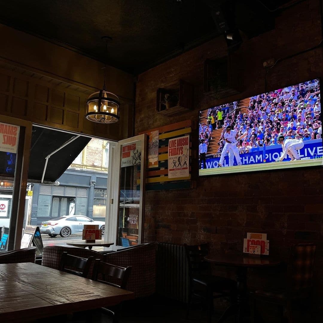 Interior of The Bower's Tap showing a cozy pub atmosphere with exposed brick walls, a wooden table, and a large TV screen displaying a live sports event. A window provides a view of the street outside.