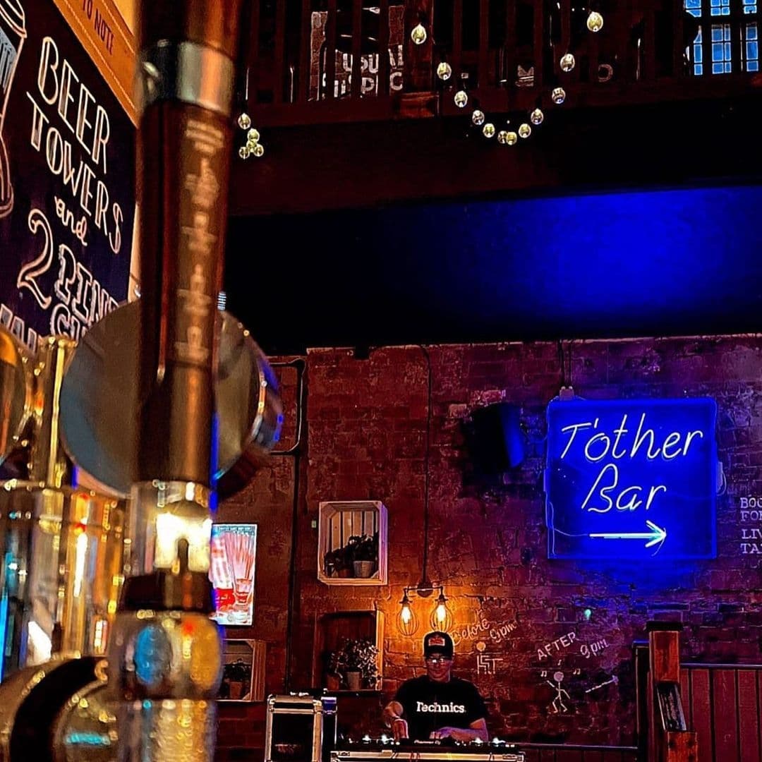 Interior view of The Bower's Tap, featuring a cozy bar atmosphere with exposed brick walls, a neon sign, and a DJ booth where a DJ is mixing tracks.