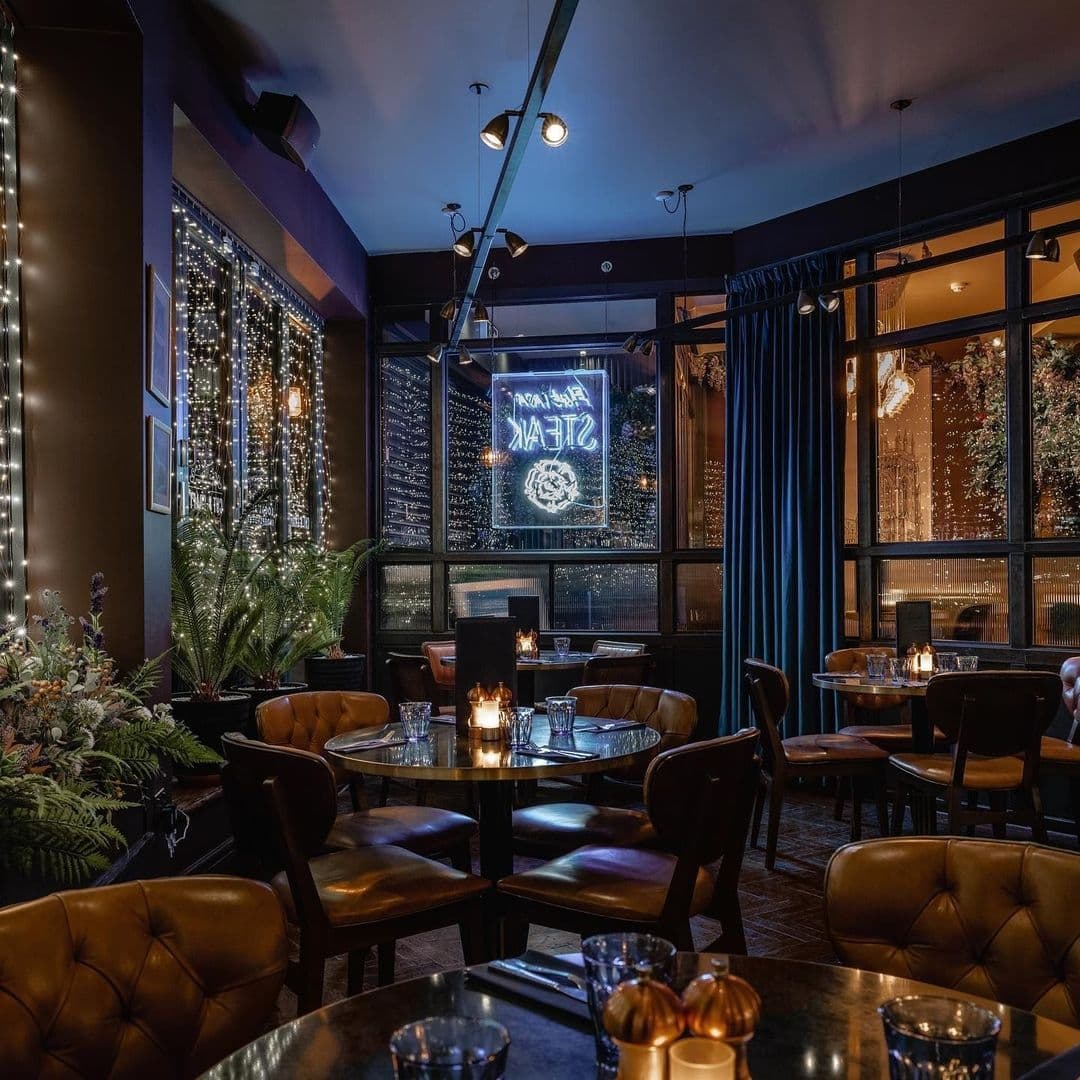 Interior of The Cut & Craft, showcasing a cozy dining area with leather chairs, round tables, ambient lighting, and decorative plants, with a neon sign in the background.