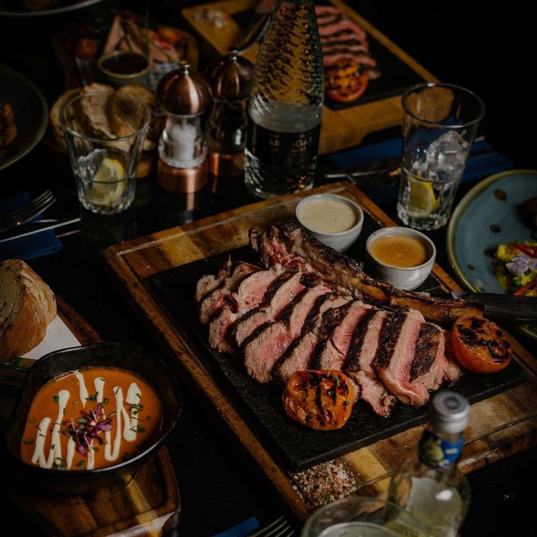 Succulent sliced steak served with grilled tomatoes on a wooden board at The Cut & Craft, accompanied by sauces, bread, and a selection of condiments.