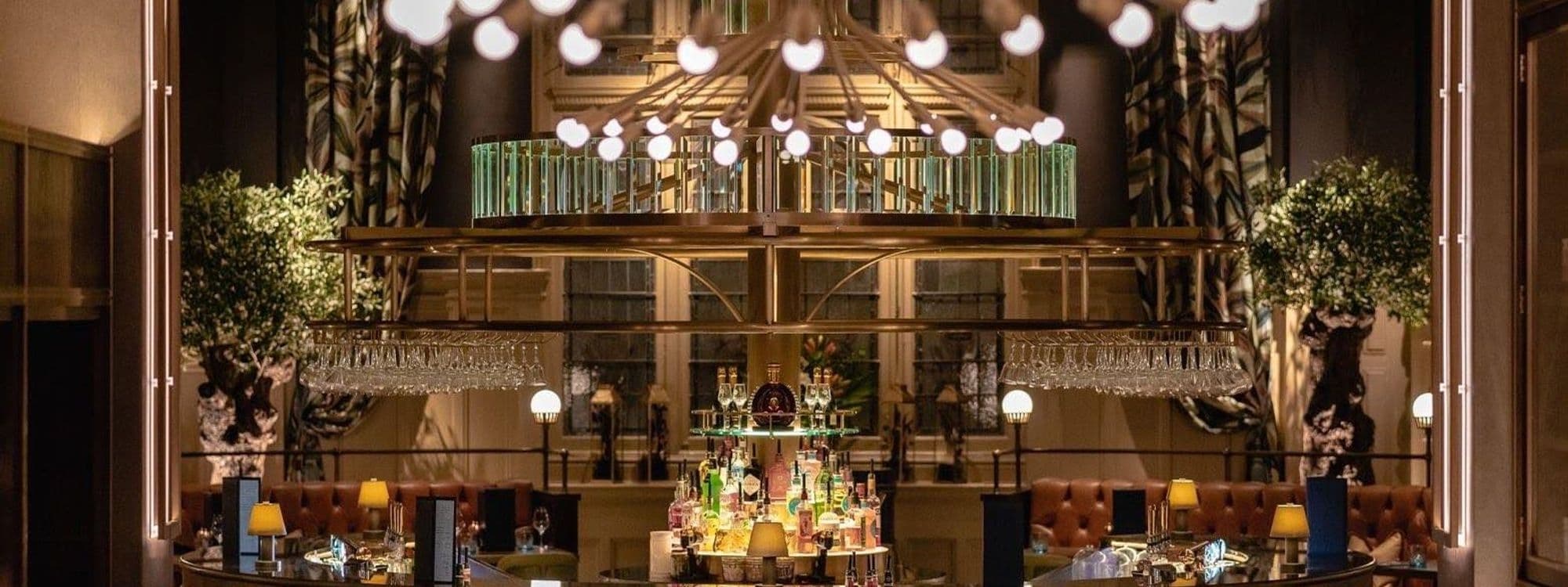 Elegant interior view of The Cut & Craft, showcasing a stylish bar with glassware, cozy seating areas with tufted leather sofas, and ambient lighting from overhead chandeliers and lamps.