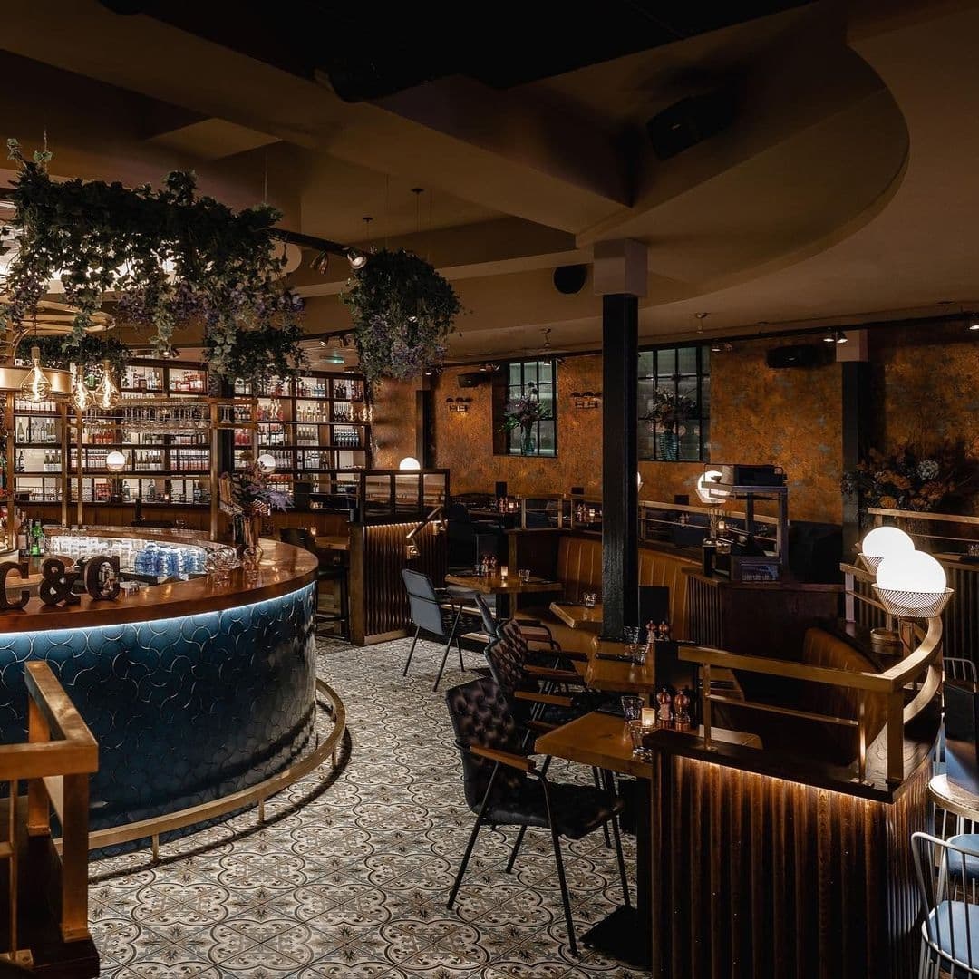 Interior of The Cut & Craft, featuring an elegant bar with blue upholstery, wooden tables, patterned floor tiles, and decorative greenery.